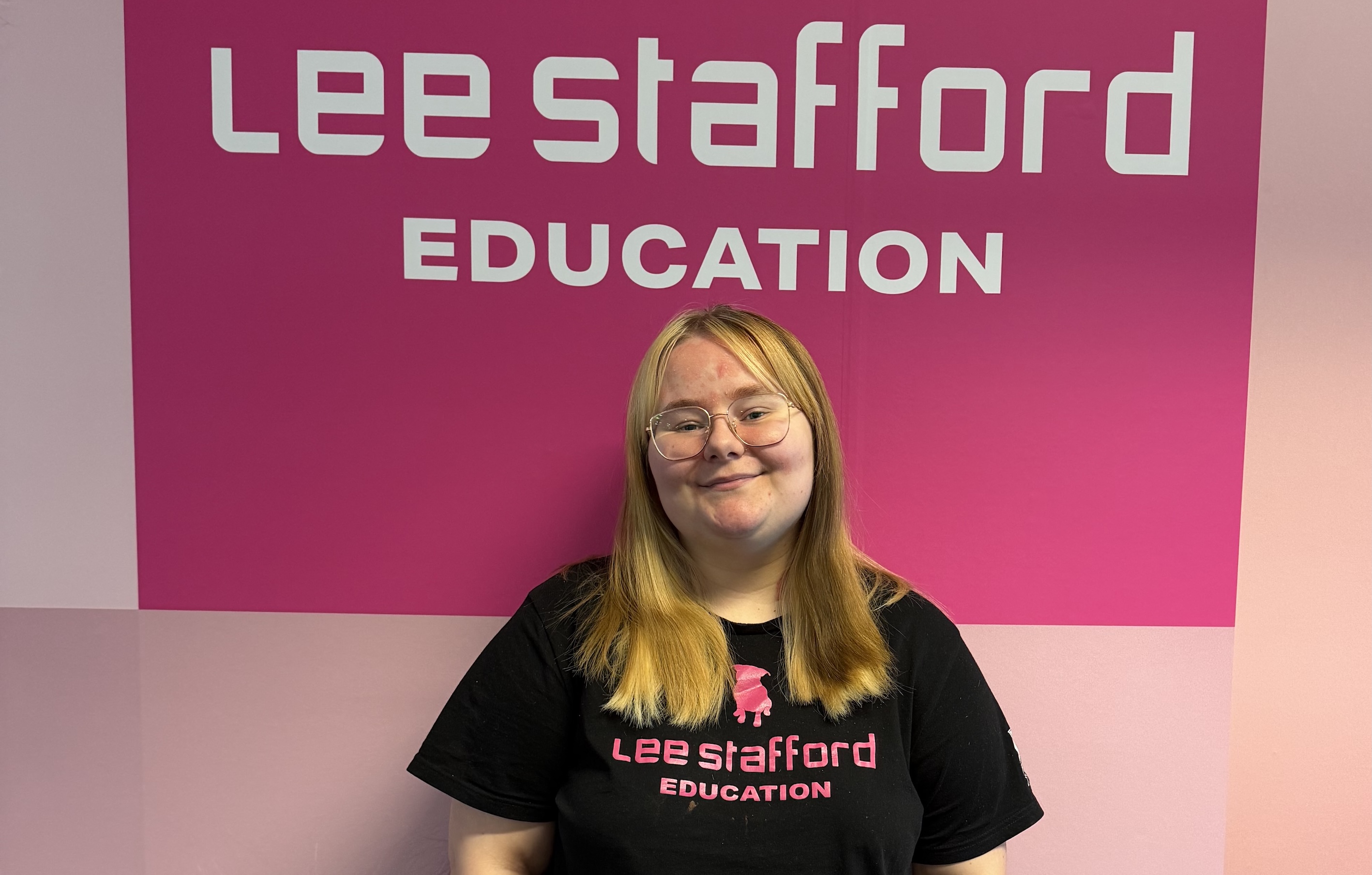 Student standing in front of pink sign saying Lee Stafford Education 